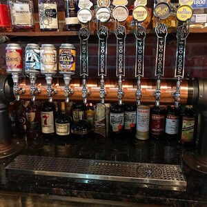 May include: A bar setup featuring multiple beer taps with "Two Roads" branding. Various liquor bottles and beer cans are visible, including "Pinner Pils" and "Zilla Pils." The taps are mounted on a copper-colored structure against a brick wall.