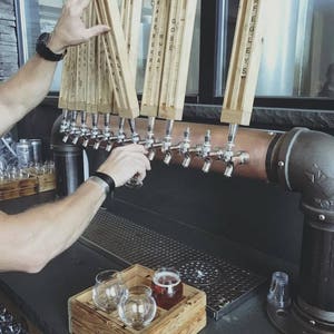 May include: A bar with beer taps and wooden tap handles. The taps are dispensing beer into glasses. A wooden tray holds two empty glasses and one filled with beer. The tap handles have text on them.