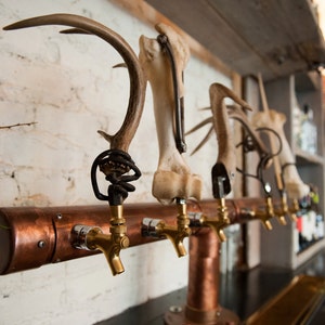 May include: A custom beer tap system with copper piping and brass taps. The tap handles are made from antlers and bones, mounted on a white brick wall. The design has a rustic, natural aesthetic.