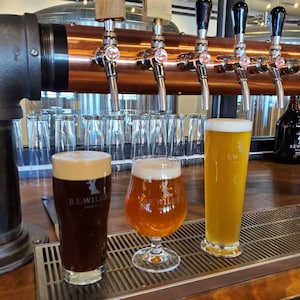 May include: Three glasses of beer with varying colors and foam levels are displayed on a bar. The beers range from dark brown to golden yellow. The text "BE WILDE" is visible on the glasses. A copper tap system is in the background.