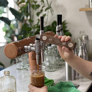 May include: A copper and stainless steel coffee tap dispensing iced coffee into a clear plastic cup. The tap has a wooden handle with printed designs. The background includes glass bottles, plants, and a green cloth.