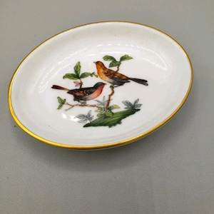 May include: An oval white ceramic dish featuring a gold-painted rim and a central illustration of two small birds perched on a green leafy branch. The birds are depicted with brown, orange, and black plumage, resting on a detailed green foliage base.
