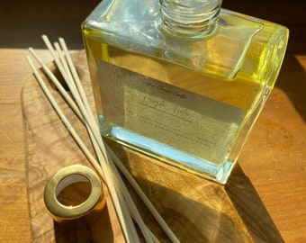 May include: A clear, square glass diffuser bottle filled with yellow liquid, with a gold-coloured cap and wooden reeds. The bottle has a label with the text "Hygge Tide". The diffuser sits on a wooden surface.