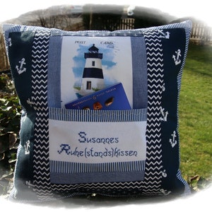 Könnte beinhalten: Ein blaues und weißes Kissen mit einer Tasche und einer Postkarte mit einem Leuchtturm darin. Das Kissen hat eine weiße Stickerei mit der Aufschrift "Susannes Ruhestandskissen".