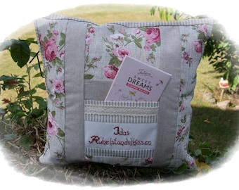 Puede incluir: Una almohada floral gris y blanca con un bolsillo en la parte delantera. El bolsillo tiene un borde de encaje y un pequeño libro en su interior. La almohada tiene el texto "Idas Ruhestandskissen" bordado en la parte delantera.