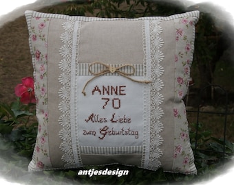 Puede incluir: Una almohada beige con un borde de encaje blanco y un estampado floral. La almohada tiene un cuadrado blanco en el centro con bordado marrón que dice "Anne 70 Alles Liebe zum Geburtstag".