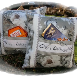 Könnte beinhalten: Zwei graue und weiße florale Kissenbezüge mit Taschen. Die Taschen enthalten Schokoladentafeln und die Kissen sind mit dem Text "Omas Lieblingsplatz" und "Uromas Lieblings" bestickt.