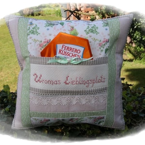 Könnte beinhalten: Ein dekoratives Kissen mit einer Tasche, mit floralen und gemusterten Stoffen in Grün-, Rosa- und Beigetönen. Die Tasche enthält eine Ferrero Rocher-Box. Der Text "Uromas Lieblingsplatz" ist auf die Tasche gestickt.