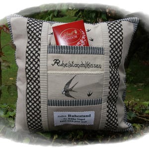 Könnte beinhalten: Ein dekoratives Kissen mit Tasche, Vogelstickerei und Text. Das Kissen hat ein Hahnentrittmuster an den Seiten und eine rote Pralinenschachtel in der Tasche. Der Text auf dem Kissen lautet "Ruhe(stands)Kissen".
