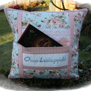 Könnte beinhalten: Ein hellblaues und rosafarbenes Blumenkissen mit einer Tasche und dem gestickten Text "Omas Lieblingsplatz". Das Kissen hat einen weißen Spitzenbesatz und ein Buch ist in die Tasche gesteckt.