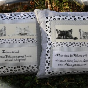 Könnte beinhalten: Zwei dekorative Kissen mit Katzenmotiven. Jedes Kissen zeigt gerahmte Katzenbilder, Pfotenabdruck-Akzente und deutschen Text. Die Kissen sind grau und weiß, mit einem schwarz-weißen Pfotenabdruck-Rand.