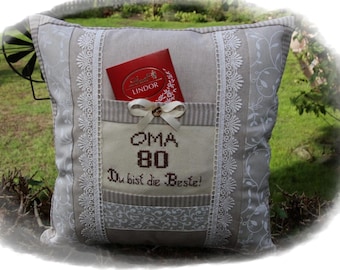 Puede incluir: Un cojín decorativo beige con un bolsillo que contiene una caja de bombones Lindor roja. El cojín tiene un ribete de encaje blanco y un diseño bordado que dice "OMA 80 Du bist die Beste!". Un lazo beige con un botón de madera adorna el bolsillo.