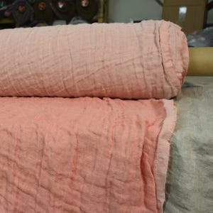 May include: Two rolls of soft, textured fabric in shades of pink and peach. The fabric appears loosely woven, with a crinkled appearance. The rolls are side by side, with the bottom roll partially unrolled, revealing the fabric's texture.