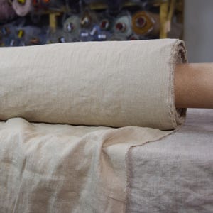May include: A roll of natural-colored linen fabric. The fabric is a light beige color and appears to be loosely woven. The edge of the fabric is frayed, and the roll is resting on a flat surface. The background shows other rolls of fabric.