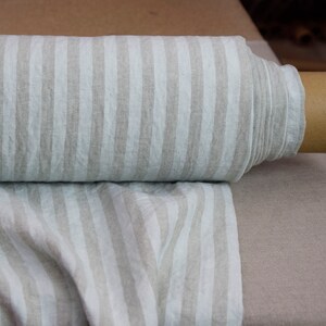 May include: A roll of fabric featuring a vertical striped pattern in alternating shades of off-white and light beige. The textile is wound around a cylindrical cardboard core, with a portion of the fabric unrolled and draped forward.