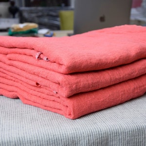 Puede incluir: Una pila de tela de lino doblada de color coral. La tela está cuidadosamente dispuesta sobre una superficie a rayas gris claro y blanco. La textura del lino es visible, lo que sugiere un material suave y natural. La pila se compone de varias capas.