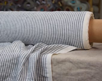 Könnte beinhalten: Eine Rolle weißen Leinenstoffs mit dünnen, dunkelgrauen Streifen. Der Stoff ist gefaltet und über eine beige Oberfläche gelegt.