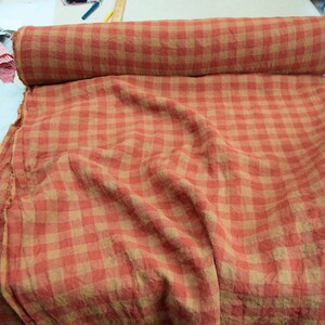 以下が含まれることがあります： オレンジとタンのチェック柄の生地のロールが白い表面に置かれています。生地は広げられており、同じパターンが表示されています。生地はリネンまたは同様の軽量素材のようで、衣類や室内装飾に適しています。