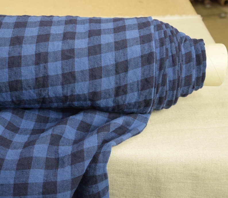 K&ouml;nnte beinhalten: Blauer und schwarzer Gingham-Stoff mit gewebter Struktur. Der Stoff ist aufgerollt und auf einer beigefarbenen Oberfl&auml;che ausgestellt.