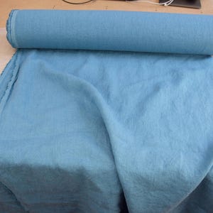 May include: A roll of light blue fabric, partially unrolled, displaying a soft, textured appearance. The fabric is a solid color, with a slight sheen. The roll is resting on a neutral-colored surface.