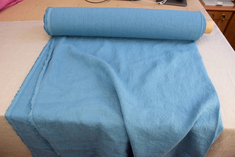 May include: A roll of light blue fabric, partially unrolled, displaying a soft, textured appearance. The fabric is a solid color, with a slight sheen. The roll is resting on a neutral-colored surface.