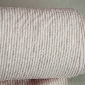 May include: A roll of natural linen fabric with thin, light brown stripes. The fabric is woven with a subtle texture and has a soft, natural feel.