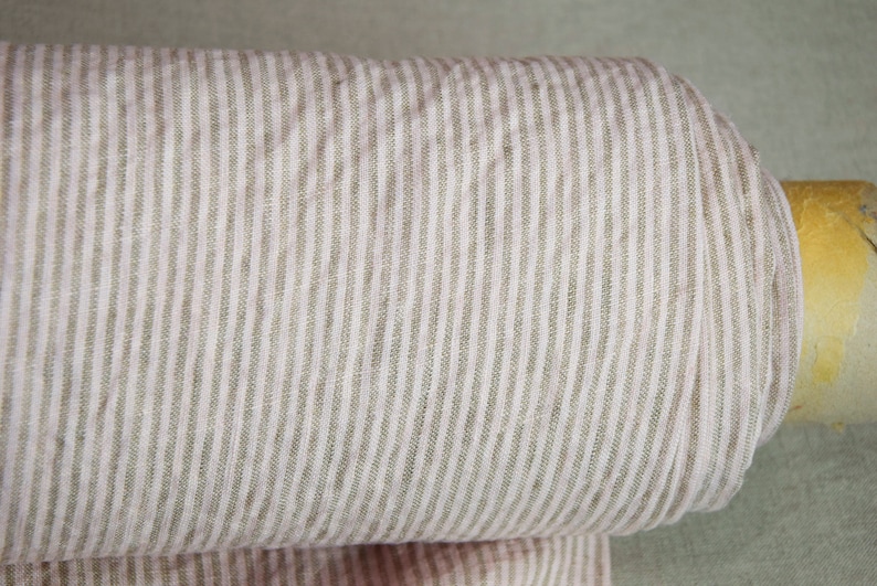 May include: A roll of natural linen fabric with thin, light brown stripes. The fabric is woven with a subtle texture and has a soft, natural feel.