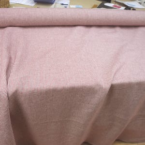 May include: A roll of red and white checkered fabric, with the fabric draped over a surface. The fabric is a light red color with a small, repeating checkered pattern in white. The fabric is rolled up at the top.