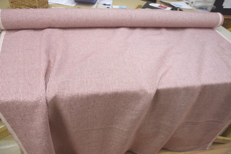 May include: A roll of red and white checkered fabric, with the fabric draped over a surface. The fabric is a light red color with a small, repeating checkered pattern in white. The fabric is rolled up at the top.