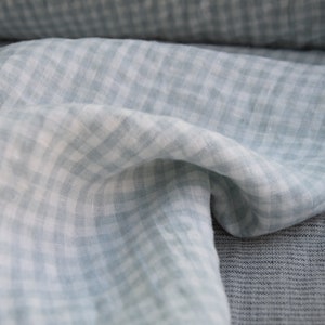 Puede incluir: Una tela de guingán azul claro y blanco con un tacto suave y texturizado. La tela está doblada y drapeada, mostrando su delicado patrón.