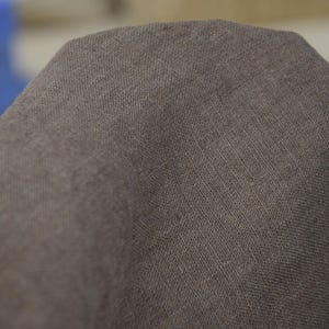 May include: Close-up of a section of brown fabric. The material has a textured appearance, with visible woven threads creating a subtle pattern. The color is a muted brown, with slight variations in tone across the surface.