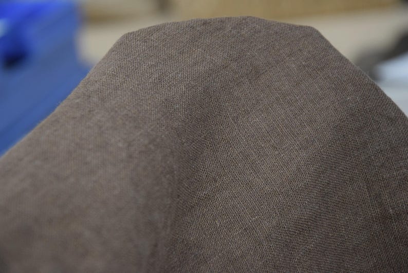May include: Close-up of a section of brown fabric. The material has a textured appearance, with visible woven threads creating a subtle pattern. The color is a muted brown, with slight variations in tone across the surface.