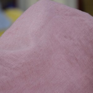 Puede incluir: Primer plano de una tela suave de color rosa empolvado. El material tiene una textura sutil y parece estar drapeado, con pliegues y sombras visibles. El color de la tela es uniforme en toda la imagen.