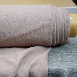 Könnte beinhalten: Eine Rolle weicher, blassrosa Stoff mit leicht strukturierter Oberfläche. Der Stoff ist aufgerollt und mit einem braunen Band gebunden.