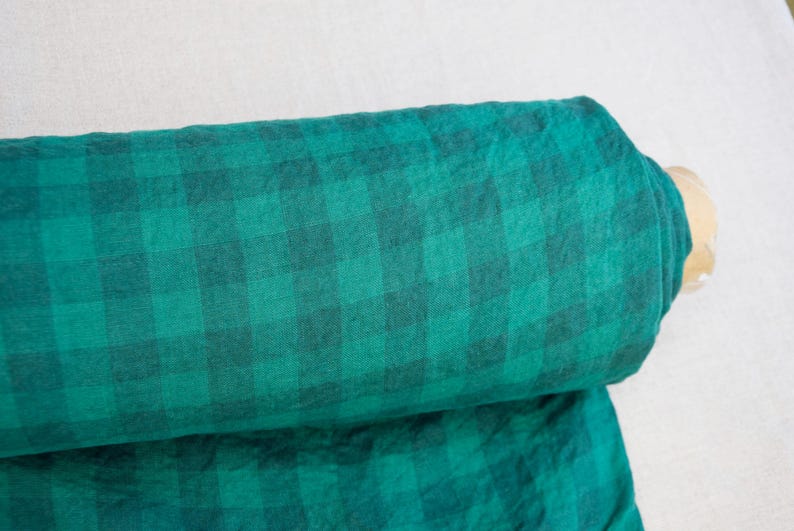 May include: A rolled-up bolt of fabric featuring a green and black plaid pattern. The fabric is a vibrant teal color with a darker green grid design. The material appears soft and possibly linen or cotton.