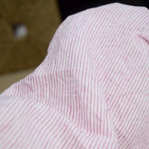 Może przedstawiać: Zbliżenie na różowo-białą tkaninę w paski. Tkanina ma miękki, teksturowany wygląd z cienkimi, pionowymi paskami. Materiał to prawdopodobnie len lub podobny lekki materiał tekstylny, odpowiedni do odzieży lub wystroju wnętrz.