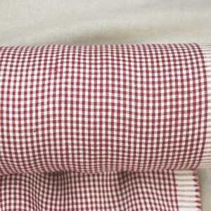 May include: Rolled-up fabric with a red and white gingham pattern. The fabric is tightly rolled, revealing a fringed edge. The gingham pattern is a classic design, with small, evenly spaced squares.