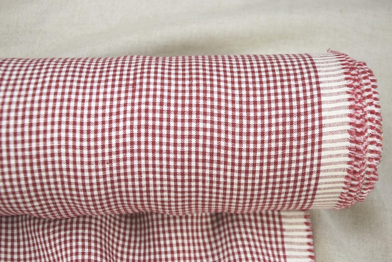 May include: Rolled-up fabric with a red and white gingham pattern. The fabric is tightly rolled, revealing a fringed edge. The gingham pattern is a classic design, with small, evenly spaced squares.
