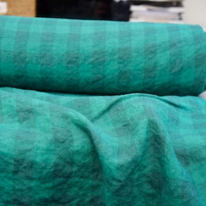 May include: A roll of green and black plaid fabric. The fabric is a deep teal color with a subtle checkered pattern. The fabric is rolled up and also draped out, showing the texture.