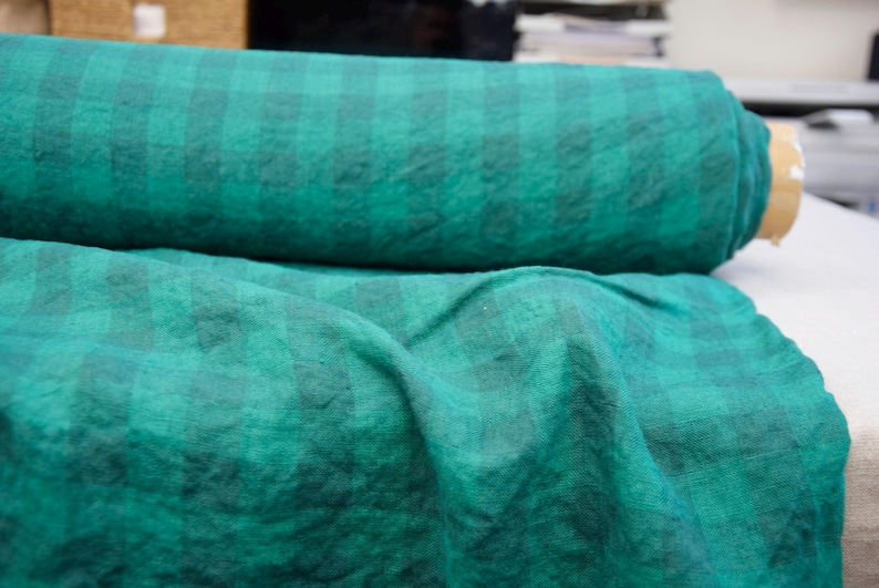 May include: A roll of green and black plaid fabric. The fabric is a deep teal color with a subtle checkered pattern. The fabric is rolled up and also draped out, showing the texture.
