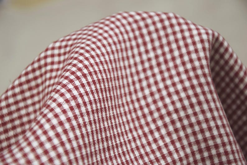 May include: Close-up of a red and white checkered fabric. The fabric is folded, showing the texture and pattern of the material. The pattern is a classic gingham design, with small squares of red and white.