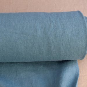 May include: A roll of teal-colored linen fabric. The fabric is tightly rolled around a cardboard tube. The fabric is a solid color and appears to be a medium weight. The background is a light tan color.