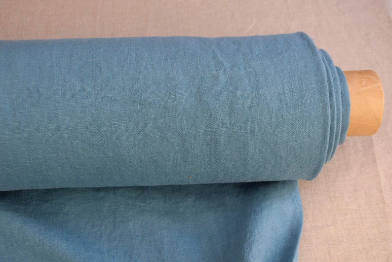 May include: A roll of teal-colored linen fabric. The fabric is tightly rolled around a cardboard tube. The fabric is a solid color and appears to be a medium weight. The background is a light tan color.