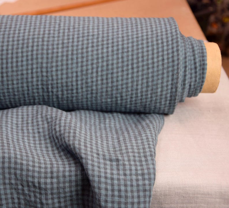 May include: A roll of blue and gray checkered fabric. The fabric is a soft, textured material, with a small gingham pattern. The roll is partially unraveled, revealing the fabric's texture and color variations. The fabric is suitable for various sewing projects.