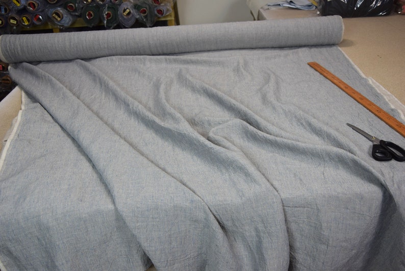 Puede incluir: Un rollo de tela gris claro con un sutil patr&oacute;n de rayas blancas. La tela est&aacute; colocada sobre una mesa con una regla de madera y unas tijeras.