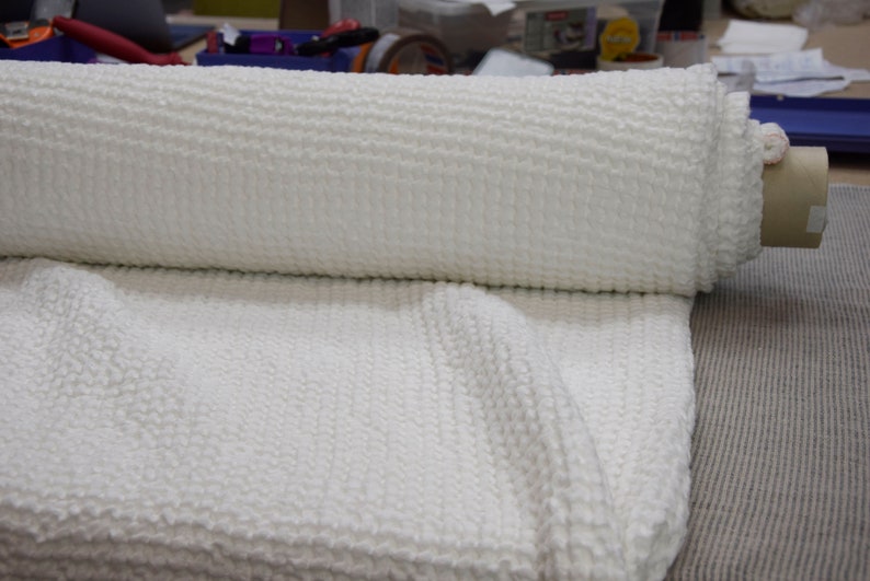 May include: A white knitted fabric rolled up on a cardboard tube. The fabric has a textured, waffle-like pattern.