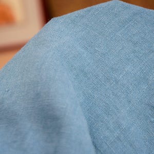 May include: Close-up of a light blue fabric with a textured weave. The fabric is folded, showing the texture and color variations. The material appears to be linen or a similar natural fiber.