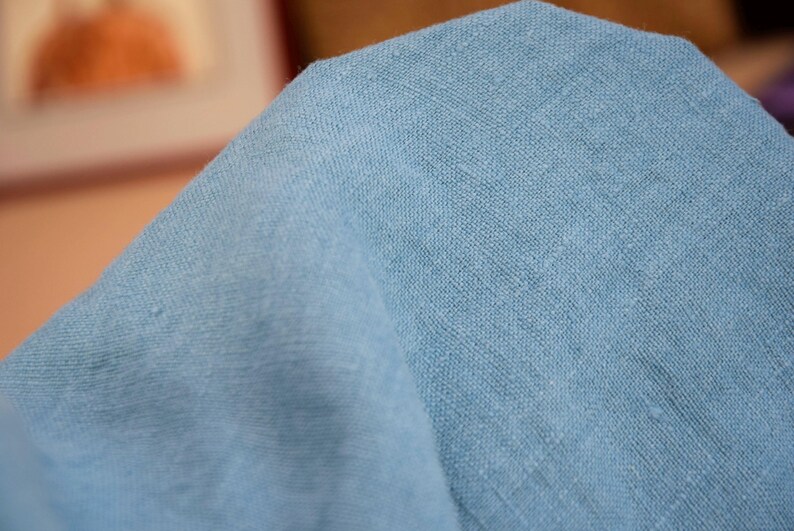 May include: Close-up of a light blue fabric with a textured weave. The fabric is folded, showing the texture and color variations. The material appears to be linen or a similar natural fiber.