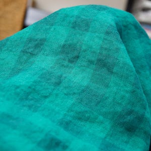 May include: Close-up of a folded piece of teal fabric with a subtle checkered pattern. The fabric has a soft, textured appearance, with variations in the teal color creating depth and visual interest. The fabric is likely used for clothing or home decor.