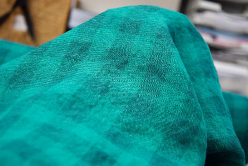 May include: Close-up of a folded piece of teal fabric with a subtle checkered pattern. The fabric has a soft, textured appearance, with variations in the teal color creating depth and visual interest. The fabric is likely used for clothing or home decor.